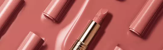 A close-up of a nude pink lipstick tube lying on a glossy pink background, surrounded by similar lipstick tubes arranged neatly.