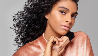 A woman with dark curly hair and a subtle glow on her face is holding a Lancome Teint Idole Ultra Wear foundation bottle, against a plain gray background.