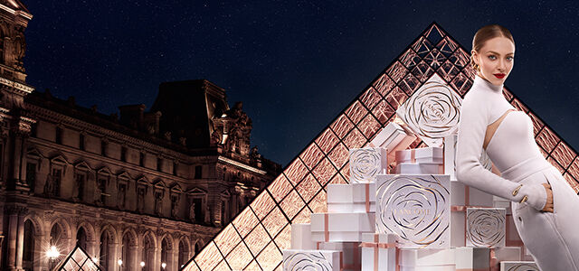 A stylish model posing with decorative gift boxes in front of the Louvre Pyramid at night, showcasing a blend of fashion and architecture.