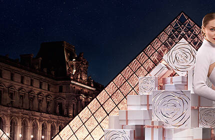 A stylish model posing with decorative gift boxes in front of the Louvre Pyramid at night, showcasing a blend of fashion and architecture.