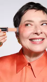 A smiling older woman with short hair holds a small makeup product up to her eye, wearing an orange blouse and gold jewelry.
