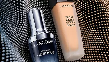 Lancome Advanced Genifique serum and Teint Idole Ultra Wear foundation are shown against a dark, textured background with rows of glowing dots.