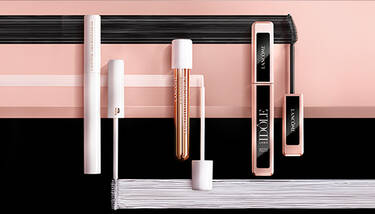Various Lancome mascaras are displayed horizontally on a pink and black background with brush strokes, showing different packaging and brush types.