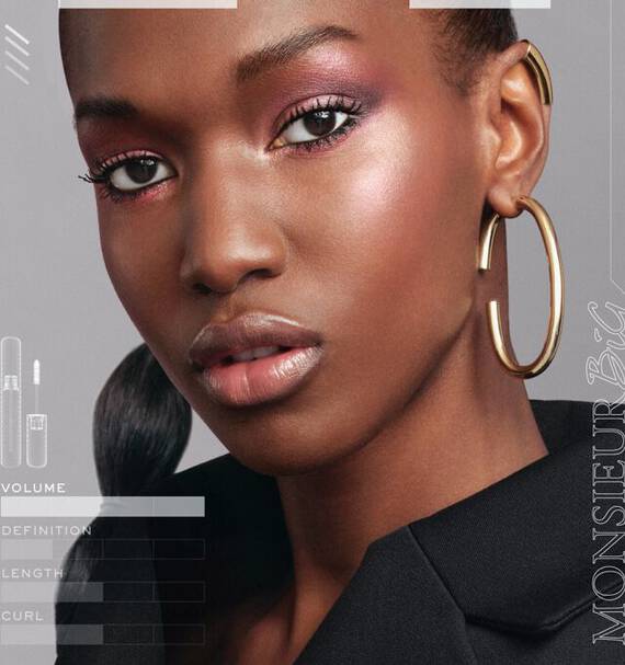 A close-up portrait of a woman with a sleek hairstyle, wearing bold makeup featuring pink tones and large hoop earrings. She is dressed in a black blazer, with elegant lighting highlighting her features. Text on the side includes makeup product details.