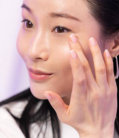 A woman gently applying skincare products to her face with a soft smile, against a light purple background.