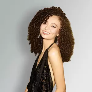 A smiling person with curly hair wearing a black outfit, posed against a light gray background.