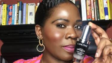 A woman with dark skin, vibrant pink lipstick, and large hoop earrings holds up a Lancome Genifique eye cream, with a blurred bookshelf in the background.