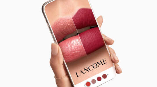 A hand holding a smartphone displaying a close-up image of lips with different shades of lipstick, featuring the Lancome logo.