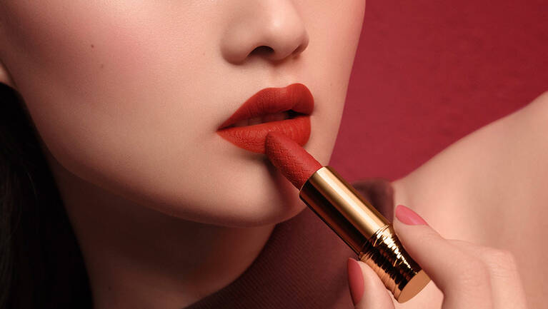 A close-up of a woman's lips being accentuated with a bold red lipstick against a rich red background.