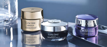 A collection of three jars of Lancome eye cream, each with distinct designs: a gold jar labeled "Absolue," a silver jar labeled "Advanced Genifique," and a purple jar labeled "Renergie Yeux Multi-Lift Ultra," arranged on a reflective surface.