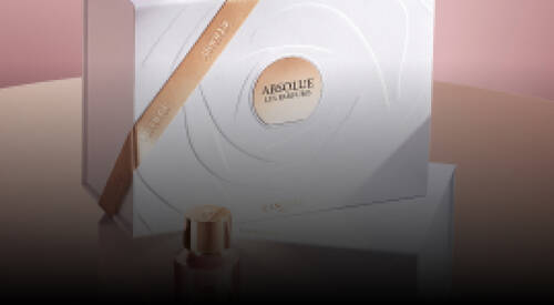 A stylish image showcasing two elegant bottles of Lancome's Absolue perfume, placed on top of a white, decorative box with subtle gold accents. The background features soft pink tones, enhancing the luxurious presentation.