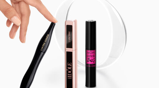 A hand pointing at three mascara products from Lancome, including a black mascara, a pink tube, and a black tube with pink lettering, arranged beside a clear geometric display.