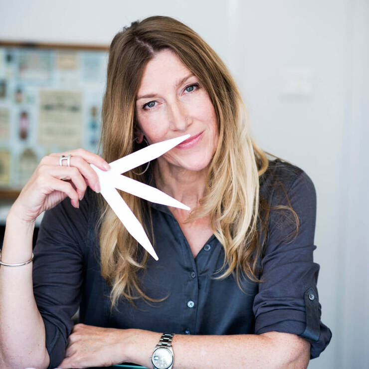 A woman with long hair playfully holds a pair of scissors near her mouth, smiling at the camera in a bright setting.