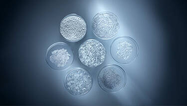 Seven petri dishes, arranged in a circle, containing various white and clear granular or flake-like substances, against a dark blue and white background.
