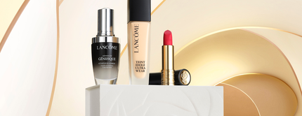 A display of Lancome beauty products, including a serum, foundation, and lipstick, arranged elegantly on a white pedestal with a soft, stylized background.