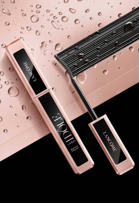 A beauty product arrangement featuring a tube of Lancome Idole Lash mascara next to a lash comb, with droplets of water on a softly textured background.