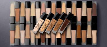 A flat lay of various foundation bottles in multiple shades, arranged in a grid pattern with some bottles prominently displayed in the center.