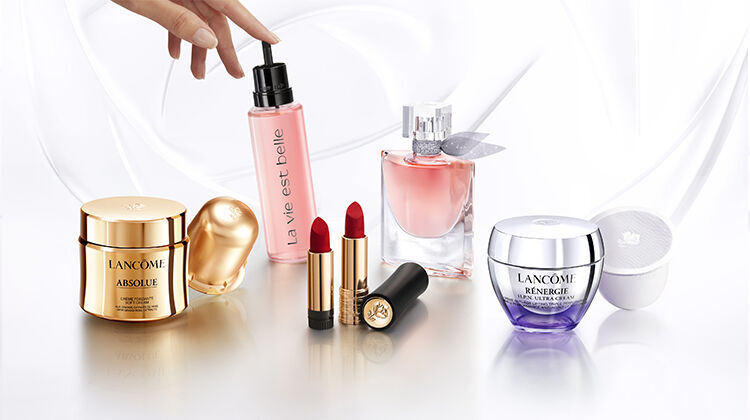 A collection of beauty products including skincare jars, lipsticks, and perfumes, arranged elegantly on a soft, light background. A hand is shown holding a pink spray bottle.