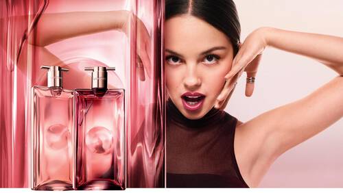 A stylish advertisement featuring two pink bottles of perfume alongside a model with long dark hair, striking a playful pose, and wearing a black top. The background is a soft gradient, enhancing the overall elegant and youthful vibe.