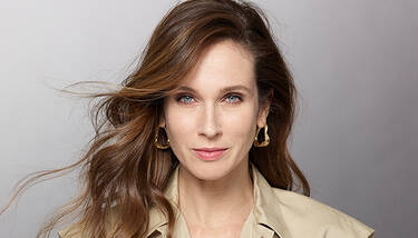A woman with blue eyes and light makeup, wearing a beige collared shirt and gold abstract earrings, looks directly at the camera with her brown hair flowing around her shoulders.
