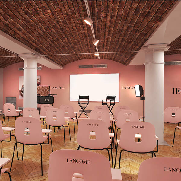 A spacious interior room designed for an event, featuring pink-colored walls and wooden flooring. Rows of white chairs with "Lancome" branding face a stage area with a black podium and large wall displays showcasing beauty imagery.