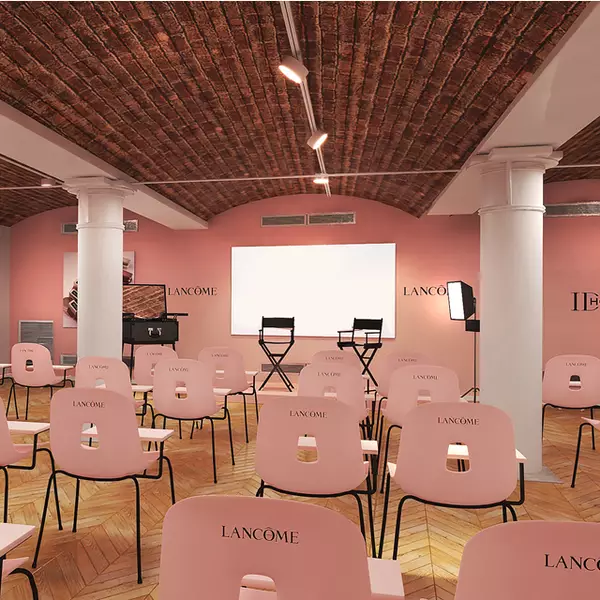 A spacious interior room designed for an event, featuring pink-colored walls and wooden flooring. Rows of white chairs with "Lancome" branding face a stage area with a black podium and large wall displays showcasing beauty imagery.