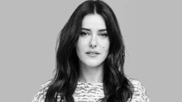 A black and white portrait of a woman with long, wavy dark hair and a serious expression, wearing a patterned top, looking directly at the camera.