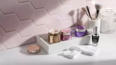 A white tray holding several Lancome skincare jars and a serum bottle, along with cotton pads and makeup brushes, sits on a counter against a pink hexagonal tiled wall.