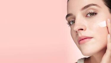 A woman with clear skin and light makeup, looking to her right, applies a small swatch of white cream to her cheek against a soft pink background.