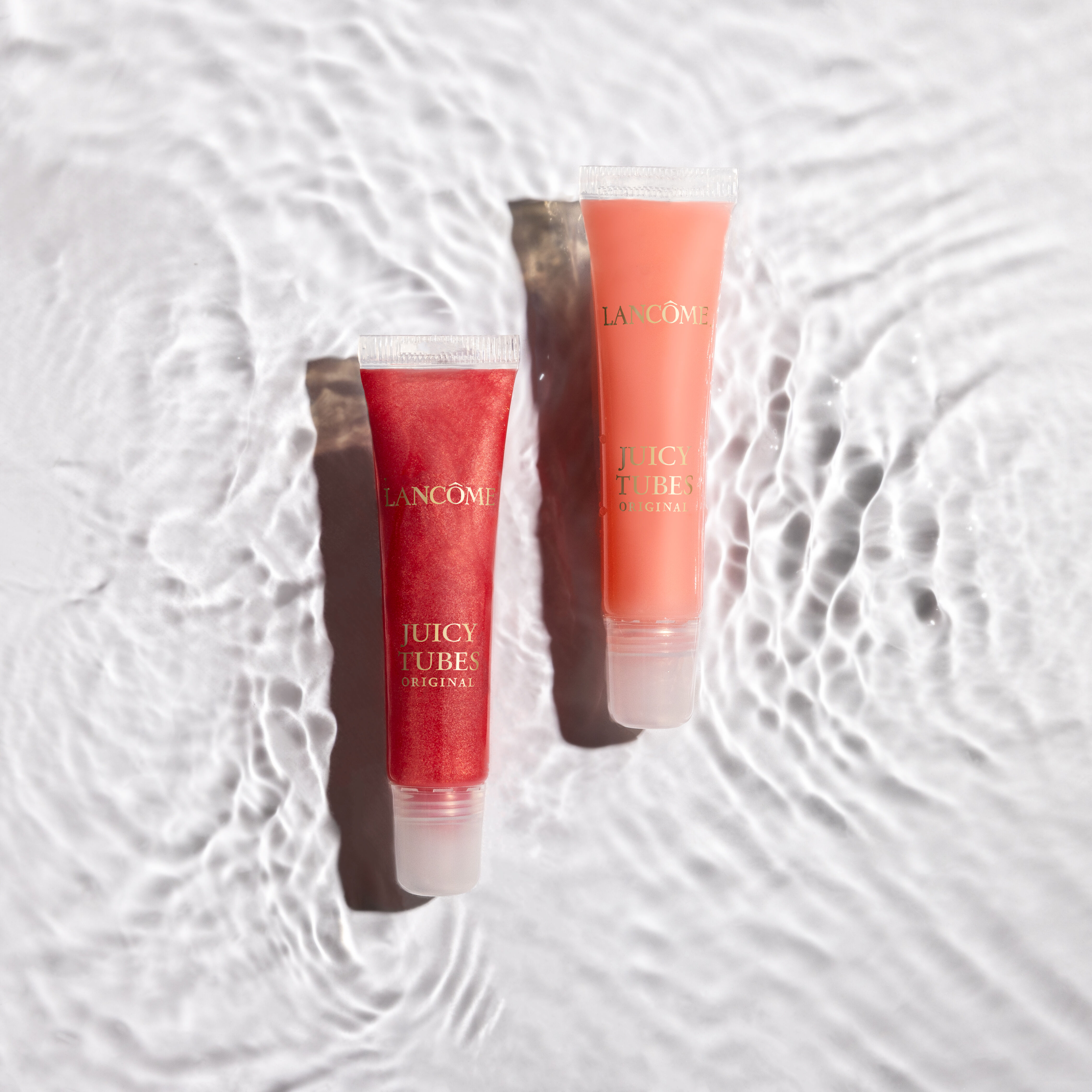 Two tubes of Lancome Juicy Tubes lip gloss sit on a textured surface that mimics rippling water. One tube contains a red gloss, and the other an orange gloss, both labeled "Juicy Tubes Original."