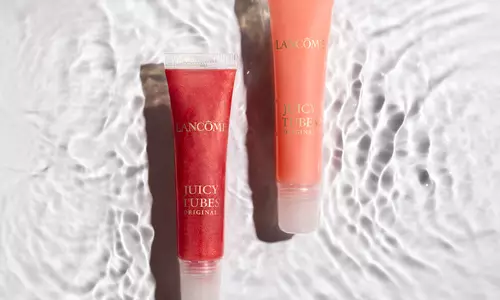 Two tubes of Lancome Juicy Tubes lip gloss sit on a textured surface that mimics rippling water. One tube contains a red gloss, and the other an orange gloss, both labeled "Juicy Tubes Original."