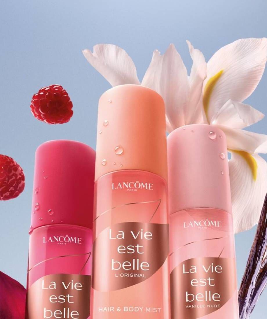 Three spray bottles of Lancome "La vie est belle" Hair & Body Mist in different shades of pink and orange, set against a background of raspberries and a white flower.