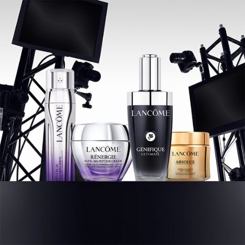 A display of Lancome skincare products, including bottles of Genifique Ultimate, Renergie Multi-Action Cream, and Absolue, arranged stylishly with a backdrop of studio lighting equipment.