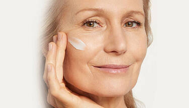 A smiling older woman applies a white cream to her cheek, looking to the right.