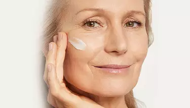 A smiling older woman applies a white cream to her cheek, looking to the right.