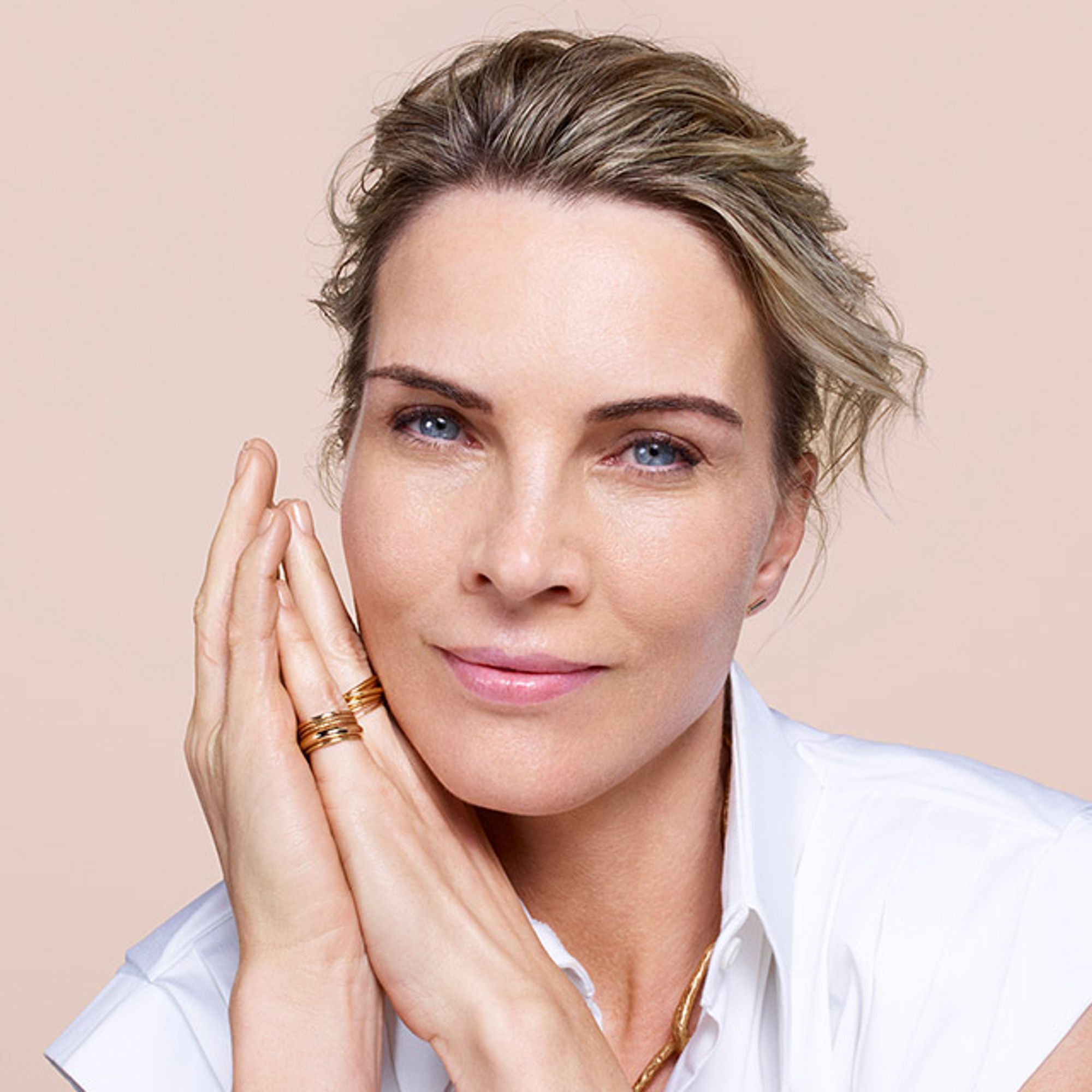 A woman with blonde hair styled back gently rests her hands on her face, smiling softly. She is wearing a white shirt, and the background features a neutral, soft color.