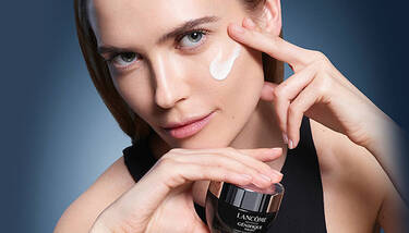 A woman applies a white cream to her cheek while holding a jar of Lancome Genifique Night cream.