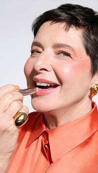A smiling woman with short hair applies lipstick while wearing a bright orange blouse and bold jewelry.