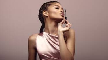 A woman in a satin top blows a kiss towards a mascara wand, with her braided hair styled back, against a neutral background.