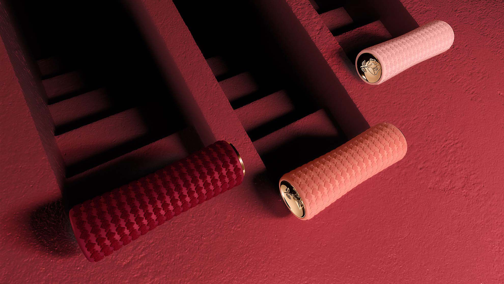 Three textured cylindrical objects in varying shades of red and pink are positioned against a textured red wall, creating a harmonious color scheme. The objects have metallic gold accents at their ends.