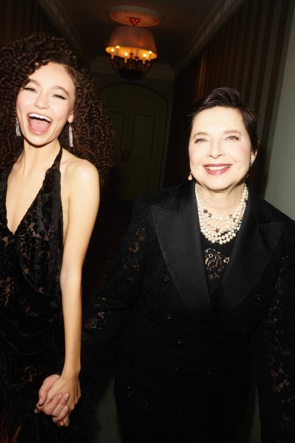 A joyful young woman with curly hair and a black dress holds hands with an elegant older woman in formal attire, both smiling brightly in a well-lit, stylish hallway.