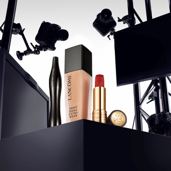 A display of Lancome makeup products, including a foundation bottle, mascara, a red lipstick, and a lipstick case, set against a backdrop of cameras and lighting equipment.