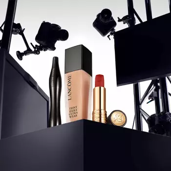 A display of Lancome makeup products, including a foundation bottle, mascara, a red lipstick, and a lipstick case, set against a backdrop of cameras and lighting equipment.