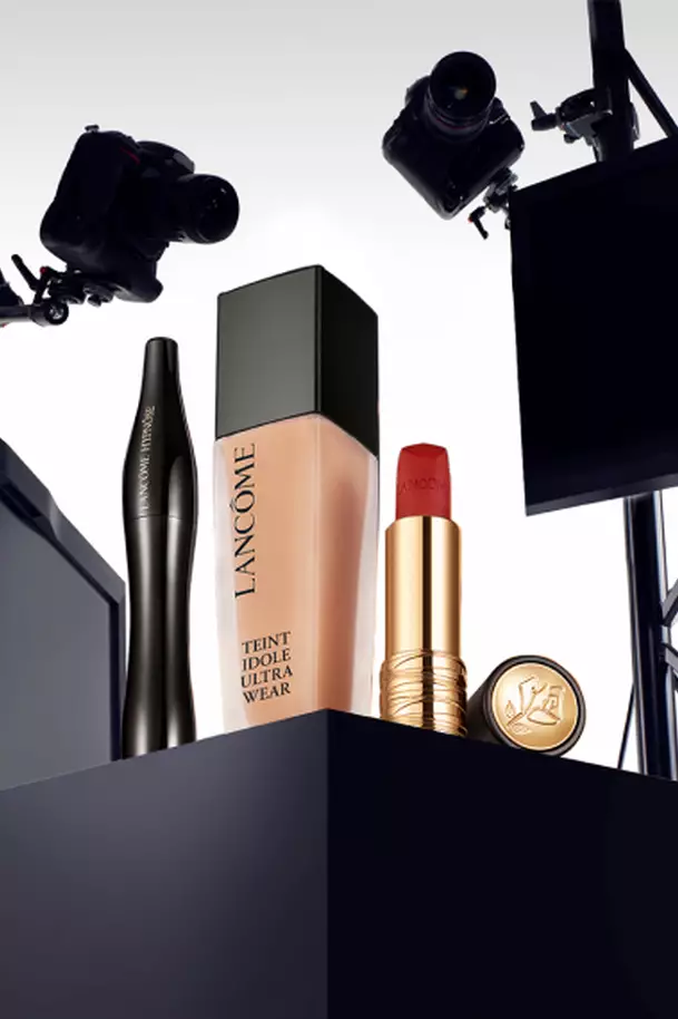 A display of Lancome cosmetics featuring a foundation bottle, a mascara, a lipstick tube, and a compact, all styled with cameras and lighting equipment in the background.