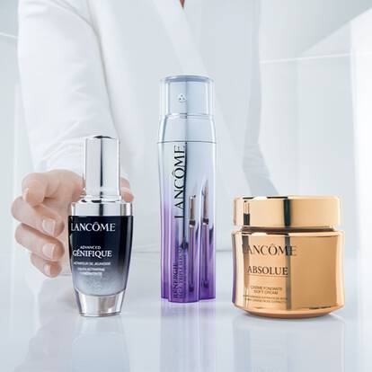 A hand reaching for three Lancome skincare products positioned on a reflective surface, featuring a serum, a treatment spray, and a moisturizer in elegantly designed containers.