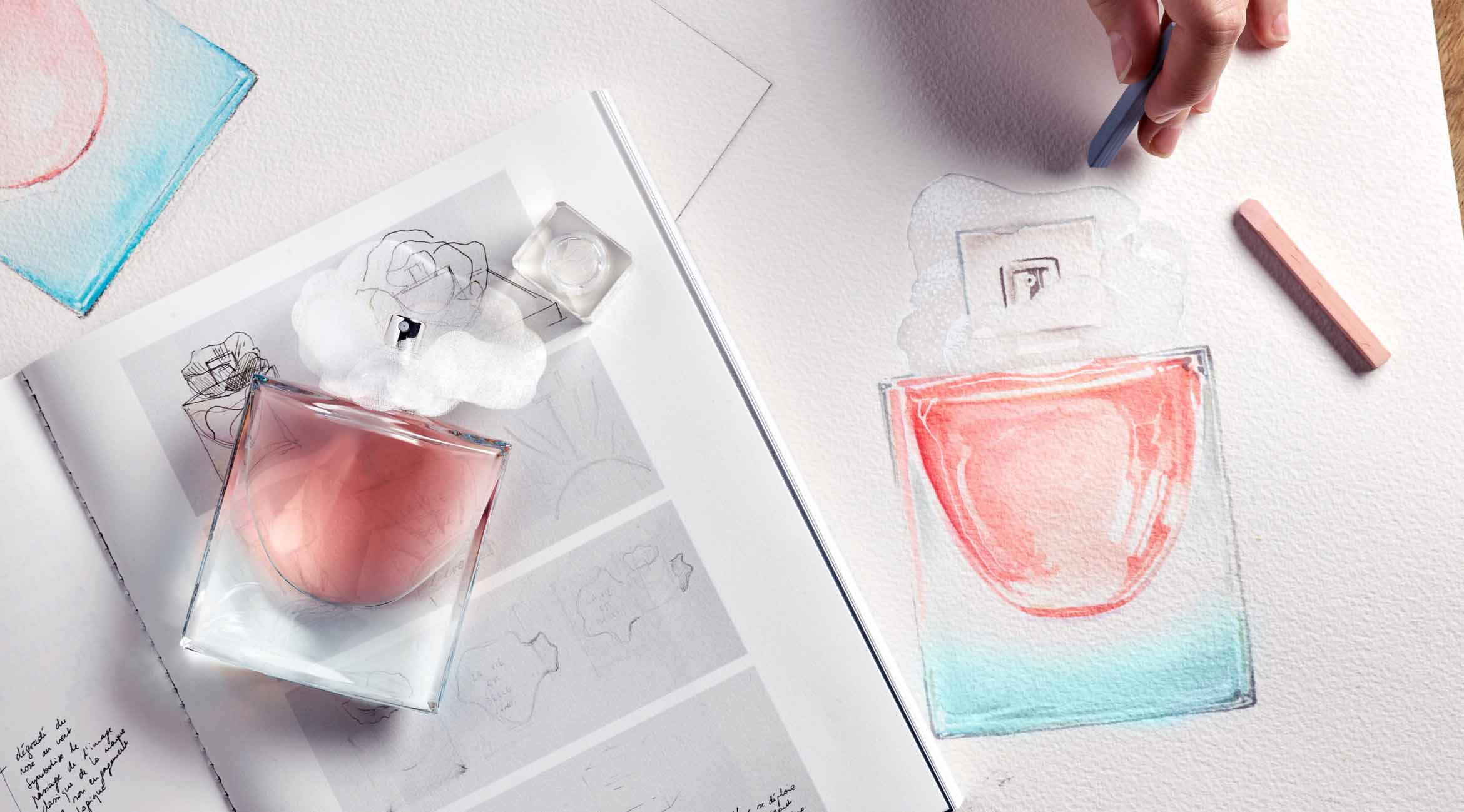 A hand colors a perfume bottle sketch while a real bottle sits beside sketches on a white artist's paper.